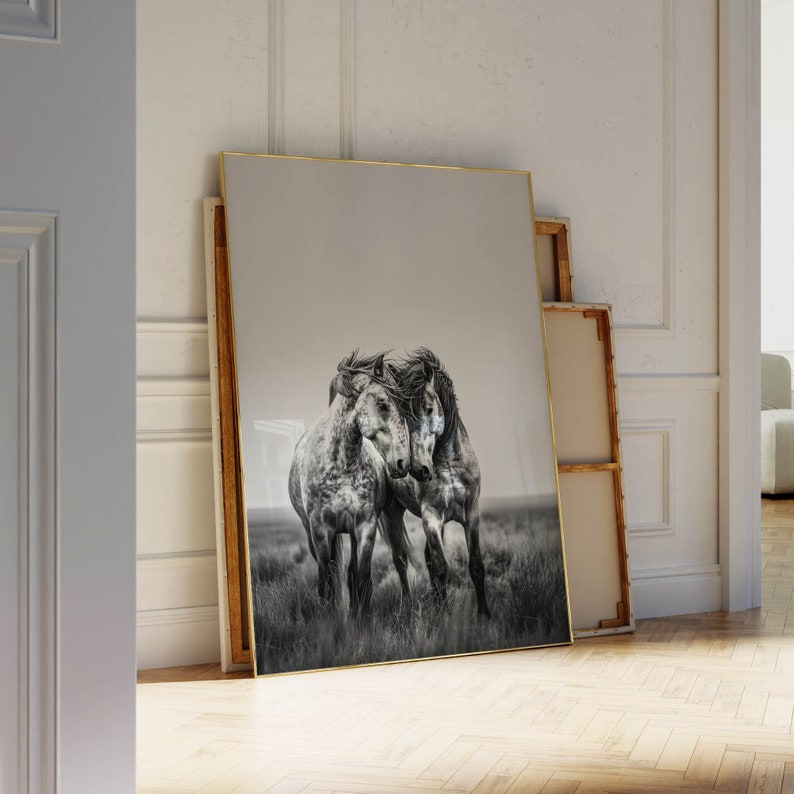 Op de afbeelding: Een zwart-witfoto van drie paarden die over een veld rennen. De paarden zijn dicht bij elkaar, met hun hoofden tegen elkaar. De foto is ingelijst in een gouden lijst.