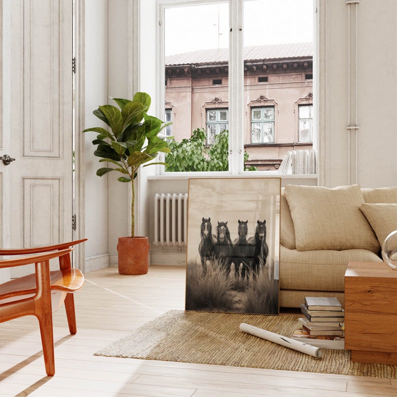 May include: A black and white photograph of four black horses running through a field of tall grass. The photograph is framed in a gold frame and is hanging on a white wall.