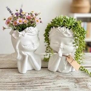 Puede incluir: Dos macetas de cerámica blanca con forma de cabeza, una con flores rosas y moradas, la otra con suculentas verdes. Las macetas tienen los ojos cerrados y un dedo en los labios, con una etiqueta en una.