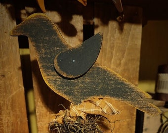 May include: A black wooden bird cutout with distressed edges, perched atop a cylindrical wooden post. The bird features a small wing detail and is accented with a textured brown fabric and dried moss nest wrapped around the base of the figure.