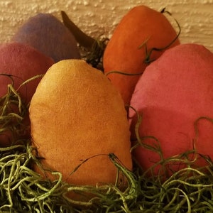 May include: A close-up of a decorative Easter display featuring five fabric eggs in shades of orange, pink, purple, and red. The eggs are nestled in a bed of green moss, creating a rustic and festive arrangement. The background is a textured, off-white surface.