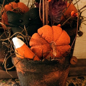 May include: A rustic Halloween decoration featuring a small burlap-wrapped pot filled with orange fabric pumpkins, a candy corn, and a black fabric bat. Twigs and berries add to the autumn theme. The pot has a decorative wire handle.