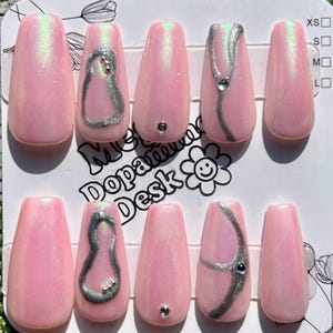 以下が含まれることがあります： ピンク色のつけ爪10本セット。パールのような仕上がり。一部の爪には、銀色の抽象的なデザインと小さなラインストーンが付いています。「Meta Dopamine Desk」の文字がパッケージに表示されています。