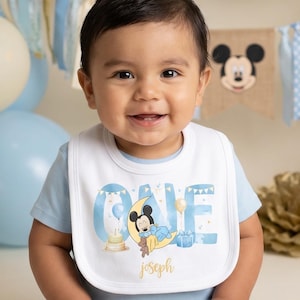 Puede incluir: Un bebé con un babero con la palabra "ONE" y un diseño de Mickey Mouse, sentado frente a un telón de fondo de globos. Un pastel con un adorno de Mickey Mouse está a la derecha. El babero también tiene el nombre "Joseph".