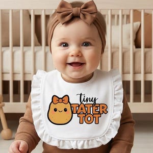 May include: A baby wearing a white bib with a ruffled edge and the words "Tiny Tater Tot" printed on it. The bib features a cartoon potato with a bow. The baby is also wearing a brown headband and a brown onesie.