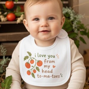 May include: A white baby bib featuring a graphic of a tomato vine with red tomatoes and green leaves. The text printed on the bib reads, "i love you from my head to-ma-toes" with a small orange heart icon between the words my and head.