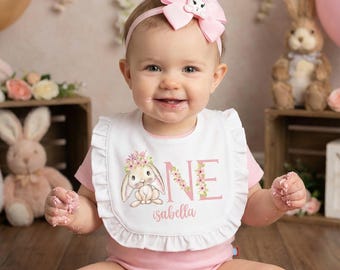Puede incluir: Un bebé con un atuendo rosa y un babero con un diseño de conejito, celebrando su primer cumpleaños. Un pastel rosa con orejas de conejo está frente al bebé. El babero dice "ONE" e "isabella". Globos y decoraciones florales en el fondo.