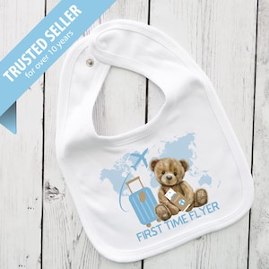 May include: White baby bib with a snap closure. The bib features a brown teddy bear with a passport, a blue suitcase, and a world map graphic. The text "FIRST TIME FLYER" is printed below the bear.