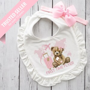Könnte beinhalten: Weißes Baby-Lätzchen mit einem rosa Schleifen-Stirnband. Das Lätzchen zeigt eine Teddybär-Illustration mit einem Koffer und einem Flugzeug sowie den Text "FIRST TIME FLYER". Das Lätzchen hat einen gerüschten Rand.