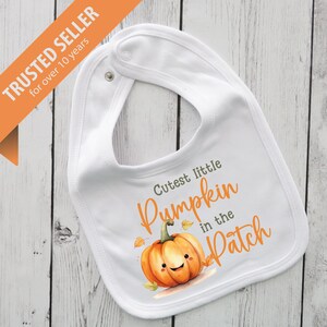 Może przedstawiać: Biały śliniaczek dla dziecka z zapięciem na zatrzask i grafiką dyni. Śliniaczek zawiera napis "Cutest little Pumpkin in the Patch" w pomarańczowym skrypcie, z ilustracją uśmiechniętej dyni.