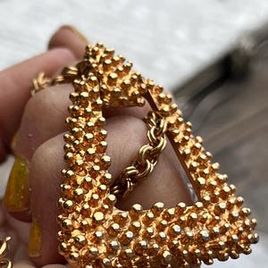 May include: Gold-toned, textured triangle earrings with a chain. The earrings have a bumpy, three-dimensional surface. The chain is a classic link style. The earrings are a bold, statement piece.