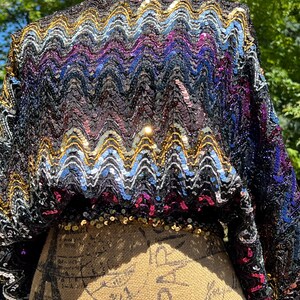 May include: A long-sleeved top covered in dense, multi-colored sequins arranged in a repeating horizontal chevron pattern. The sequins feature shades of black, gold, silver, blue, and magenta, creating a vibrant, reflective surface across the garment.