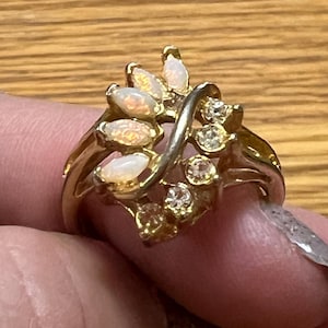 May include: A gold-toned ring featuring a cluster of oval-shaped opals and small, round clear gemstones. The ring has a leaf-like design with a curved band. The opals have a milky, iridescent appearance.