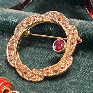 May include: A circular gold-tone brooch featuring a textured, braided-style border set with small clear stones. A single, round, deep red gemstone is bezel-set on the inner edge of the circle. A thin metal pin mechanism is visible across the center.