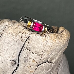 May include: A silver ring with a rectangular, bright pink gemstone. The ring has gold accents on either side of the gemstone and a textured band. The ring is displayed on a piece of weathered wood.