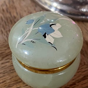 May include: A small, round, pale green stone trinket box with a polished finish. The lid features a painted floral design in blue, white, and black, with a signature. A thin, shiny gold-colored metal band separates the lid from the base.