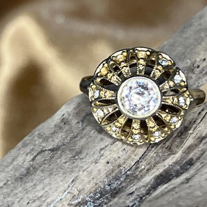 May include: Close-up of a vintage-style ring with a large, clear, round center stone. The ring features a gold-toned filigree design with small, sparkling accent stones. The band is dark, and the ring is set against a textured, wooden surface.