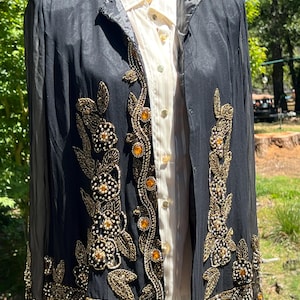 May include: A black long-sleeved jacket featuring intricate gold beadwork and embroidery in floral patterns along the front panels, collar, and cuffs. The jacket is layered over a white button-down shirt with a subtle vertical striped texture.