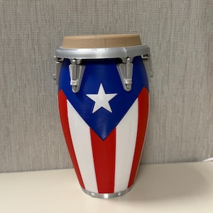 May include: A conga drum featuring the Puerto Rican flag design. The drum has a blue top with a white star, and red and white stripes on the body. The drum's rim is silver, and the top is tan.