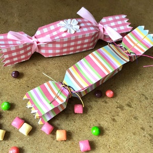 May include: Two colorful paper party favors in the shape of candy canes. The favors are tied with pink and multi-colored string. The favors are surrounded by small, colorful candies.