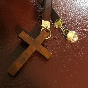Könnte beinhalten: Ein brauner Holz-Kreuzanhänger mit einer goldenen Kette und einem kleinen Glasglockenanhänger. Der Anhänger hängt an einem braunen Lederband.