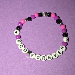 May include: A beaded bracelet featuring alternating black, purple, and bright pink round beads, interspersed with silver-toned accents. The bracelet includes white letter beads spelling out the text PCD FOREVER and two white heart-shaped beads.