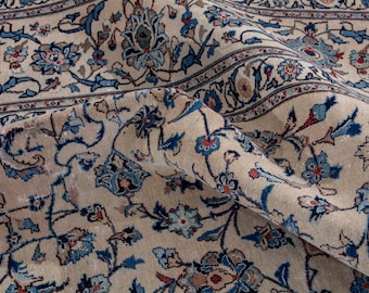 May include: Close-up of a beige rug with an intricate floral pattern in shades of blue, red, and beige. The rug has a detailed border and a folded section, showcasing the texture and design of the oriental rug.