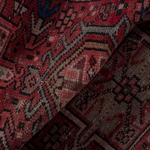 May include: Close-up of a folded, patterned rug with a rich, red background. The rug features geometric designs in black, gray, and beige, creating a textured appearance. The intricate details and color palette suggest a vintage or traditional style.