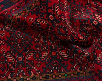May include: Close-up of a red, patterned rug with a traditional design. The rug features a repeating geometric pattern in navy blue, brown, and white. The rug's texture is visible, with slight folds and variations in color.