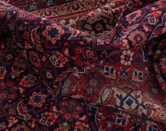 May include: Close-up of a rolled-up, intricately patterned rug. The rug features a rich color palette of deep red, navy blue, and beige. Geometric and floral designs are visible throughout the rug's surface, creating a traditional aesthetic.