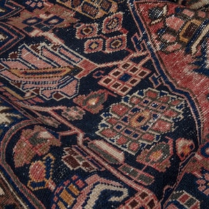 May include: Close-up of a folded, antique rug with a detailed, geometric pattern. The rug features a dark navy blue background with intricate designs in shades of red, beige, and brown. The texture appears soft and worn.