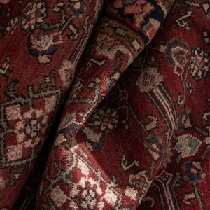 May include: A deep red area rug featuring a dense, intricate pattern of geometric and floral motifs in shades of navy blue, cream, and muted sage green. The rug is folded over, revealing the textured pile and the complex, repeating design elements.