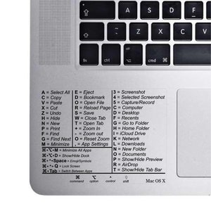 May include: A close-up of a silver laptop keyboard with a white sticker showing keyboard shortcuts for a Mac computer. The sticker lists keyboard shortcuts for common actions like Select All, Copy, Paste, Cut, Undo, Hide, New, Print, Find, Find Next, Minimize, Eject, Bookmark, Open File, Reload Page, Save, Close Tab, Open Tab, Zoom In, Zoom Out, Reset Zoom, App Settings, Screenshot, Selected Screenshot, Capture/Record, Computer, Desktop, Recents, Go to Folder, Home Folder, iCloud Drive, Network, Downloads, New Folder, Documents, Show/Hide Preview, AirDrop, and Show/Hide Tab Bar. The sticker also includes the text "Mac OS X".