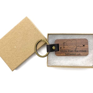 May include: A wooden keychain with a brown leather strap and a brass key ring. The keychain has a heart shape with the words "I love you" and a plane and pin graphic. The text below the heart reads "More than the miles between us."