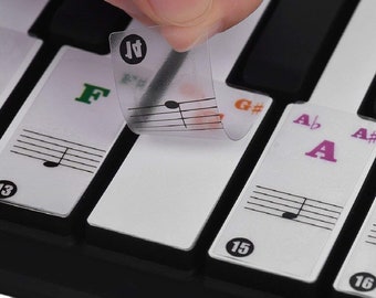 May include: A close-up of a keyboard with clear stickers that have musical notes and letter names on them. The stickers are being applied to the keys. The keys are labeled with numbers, letters, and sharps. The stickers are labeled with the letters A, F, G, and A#.