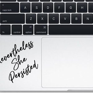 May include: A laptop computer with a black and white sticker on the trackpad that says "Nevertheless She Persisted."