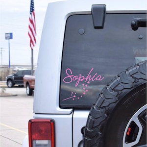May include: A pink vinyl decal with the name "Sophia" in cursive script and a pink magic wand with stars. The decal is on the back window of a silver car.