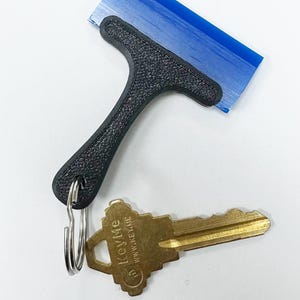 May include: A black, T-shaped squeegee with a blue blade sits above a gold-colored key on a white surface. The key is attached to a silver key ring. The key has the text "KeyMe" printed on it.