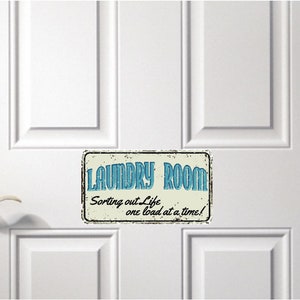 May include: A vintage-style sign with a distressed white background and black border. The sign reads "Laundry Room" in blue text with the words "Sorting out Life one load at a time!" in smaller blue text below.