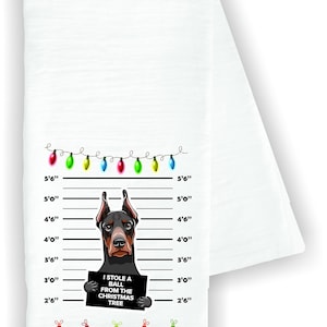 May include: White kitchen towel featuring a Doberman Pinscher mugshot design. The dog holds a sign that reads "I STOLE A BALL FROM THE CHRISTMAS TREE." The towel includes a string of Christmas lights and decorative ornaments.