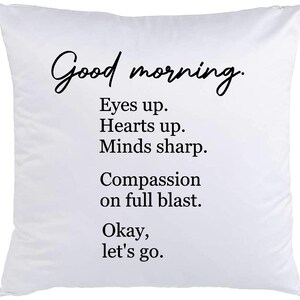 May include: White throw pillow with black text that reads "Good morning. Eyes up. Hearts up. Minds sharp. Compassion on full blast. Okay, let's go."