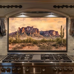 May include: A framed landscape painting of a desert scene with a mountain range and cacti. The artwork is displayed above a stainless steel gas range in a kitchen setting. The sky is a warm orange and yellow.