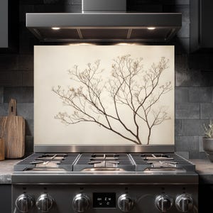 Puede incluir: Un protector contra salpicaduras de cocina decorativo con una imagen en tonos sepia de una silueta de rama de árbol. La imagen está montada detrás de una estufa de gas de acero inoxidable, con una pared de azulejos negros en el fondo.