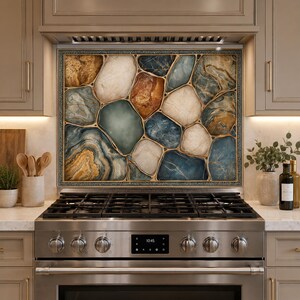 May include: A rectangular kitchen backsplash panel featuring a mosaic pattern of irregular, stone-like shapes in shades of blue, cream, tan, and brown, outlined with thin gold lines, positioned behind a stainless steel gas stove and oven range.