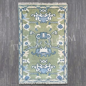 May include: A rectangular area rug featuring a sage green background with symmetrical patterns in light blue, white, and cream. The design includes stylised floral and geometric motifs, a decorative border, and short fringe along the top and bottom edges.