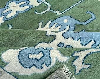 Könnte beinhalten: Ein grüner Teppich mit einem sich wiederholenden Muster aus hellblauen und weißen abstrakten Formen. Der Teppich hat eine weiche Textur und einen Fransenrand. Das Design ist auf den gefalteten Abschnitten des Teppichs sichtbar.