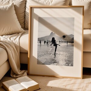 May include: Framed black and white photograph of a person kicking a ball on a beach. The image is set against a backdrop of mountains and a cloudy sky, with other people in the distance.