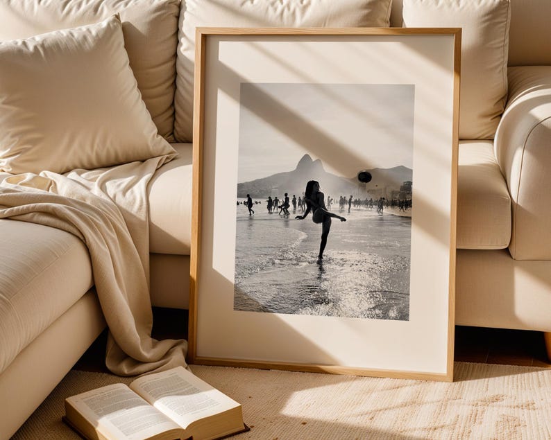 May include: Framed black and white photograph of a person kicking a ball on a beach. The image is set against a backdrop of mountains and a cloudy sky, with other people in the distance.