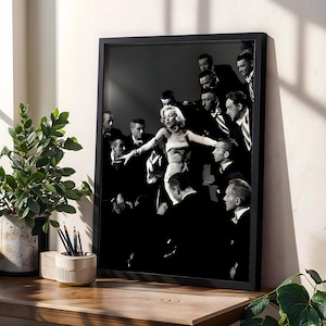 May include: Black and white framed print featuring a vintage photograph of Marilyn Monroe, surrounded by men in suits. The image is displayed on a wooden surface, next to a potted plant and a pencil holder.
