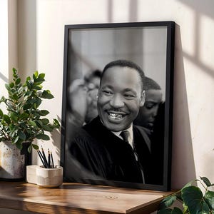 Puede incluir: Retrato enmarcado en blanco y negro de Martin Luther King Jr. sonriendo. La fotografía está en un marco negro y se muestra sobre una superficie de madera. La imagen está junto a una planta en maceta y un portalápices.